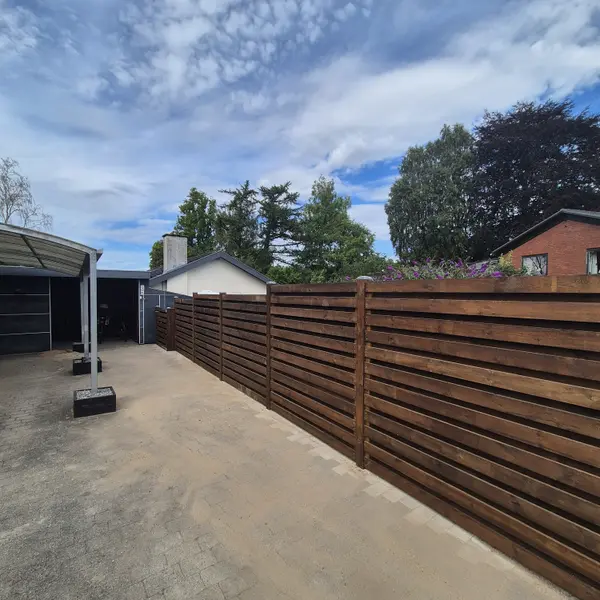 Langt træhegn i vandrette brædder langs asfalteret gårdplads