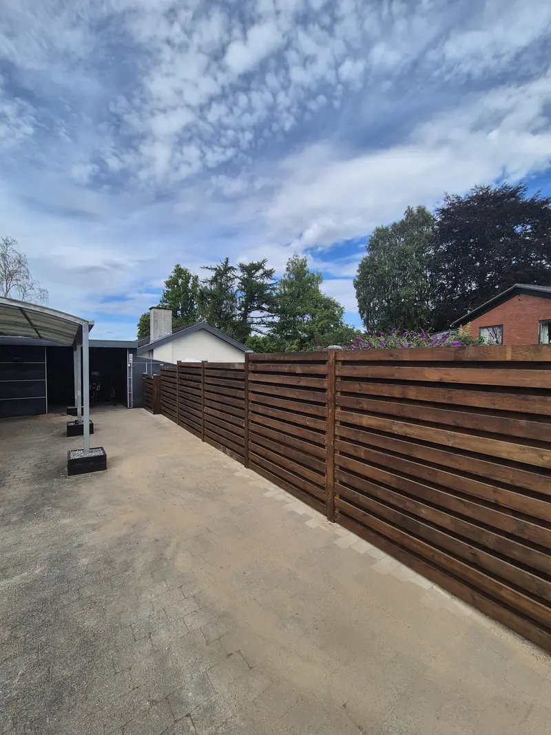 Langt træhegn i vandrette brædder langs asfalteret gårdplads